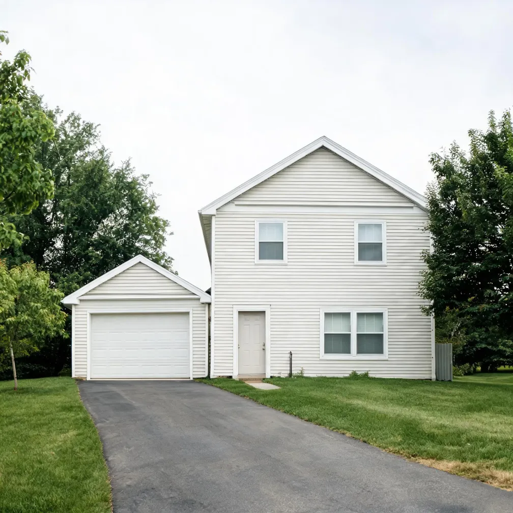 A two-story house with white horizontal vinyl siding and an attached two-car garage, fronted by an asphalt driveway, mown lawn, and deciduous trees under an overcast sky.