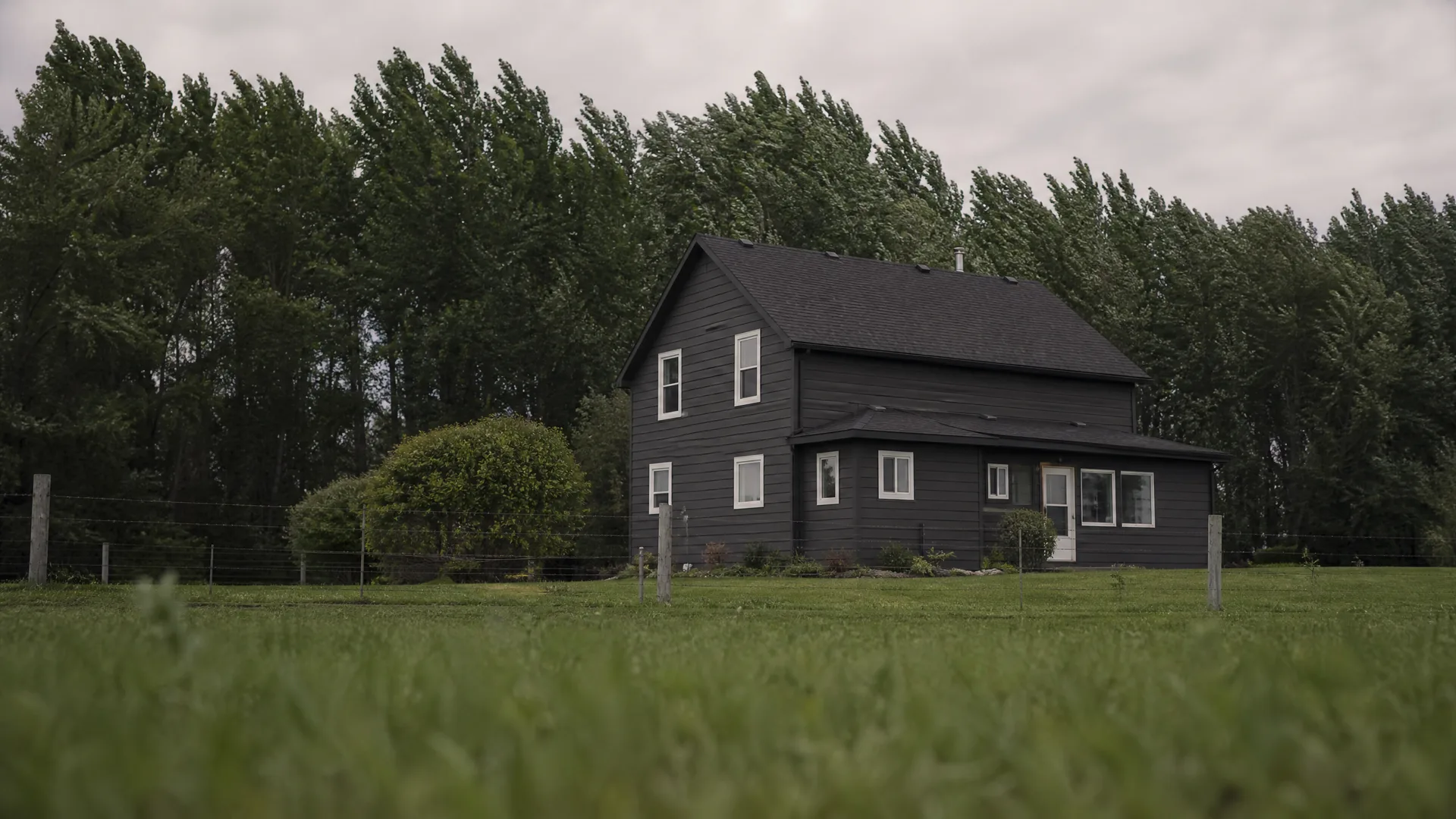 A dark-painted two-story house with white-trimmed windows and a gray shingled roof stands behind a wire fence in a grassy field, backed by tall deciduous trees under an overcast sky.