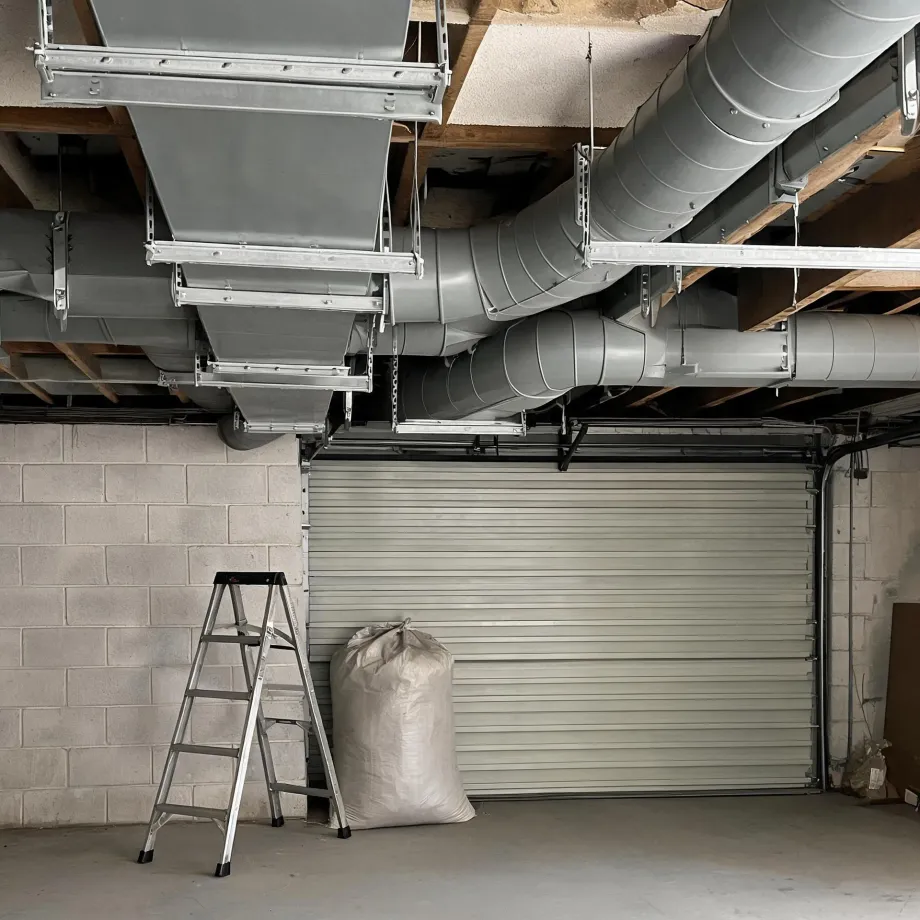 Interior of a garage with concrete block walls and a closed roll-up door, with round and rectangular metal ventilation ducts suspended from the wooden ceiling joists, a folding step ladder and a filled woven sack on the floor.