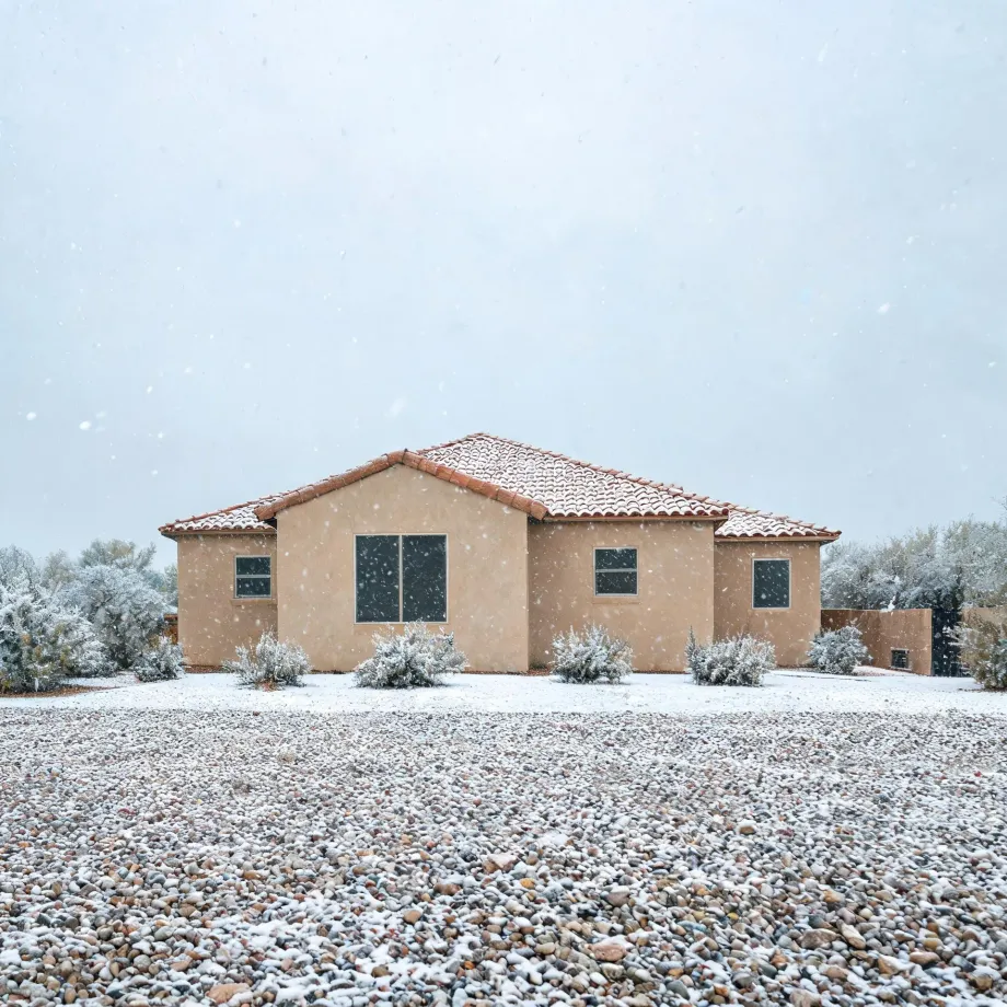 A single-story tan stucco house with a red tile roof during falling snow, with low snow-dusted shrubs and a broad gravel yard in the foreground.