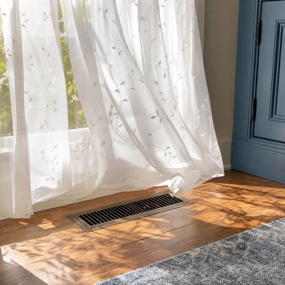 A metal floor vent set into hardwood flooring near a window with sheer leaf-patterned curtains, with sunlight casting leaf shadows and a gray rug at lower right.