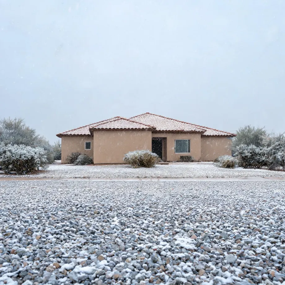 A single-story tan stucco house with a tile roof, surrounded by low shrubs and a gravel yard, all lightly covered in snow under an overcast sky.