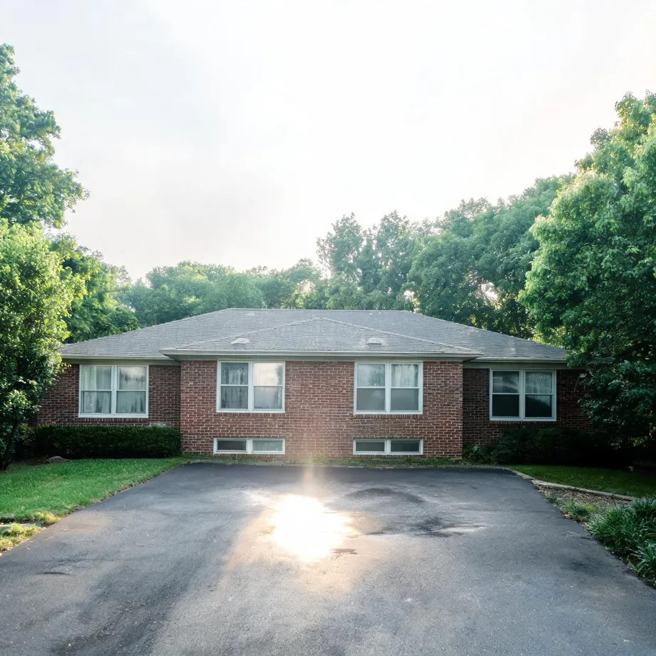 A single-story brick ranch house with a gray shingled hip roof, white-framed windows and basement windows, fronted by a wide asphalt driveway with sun glare, surrounded by green lawn and leafy trees under a bright hazy sky.