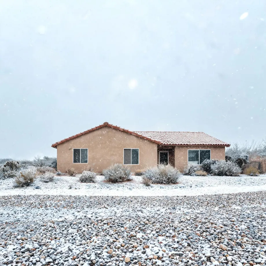A single-story tan stucco house with a red tile roof during falling snow, surrounded by snow-dusted desert shrubs and a gravel yard in the foreground under a pale overcast sky.