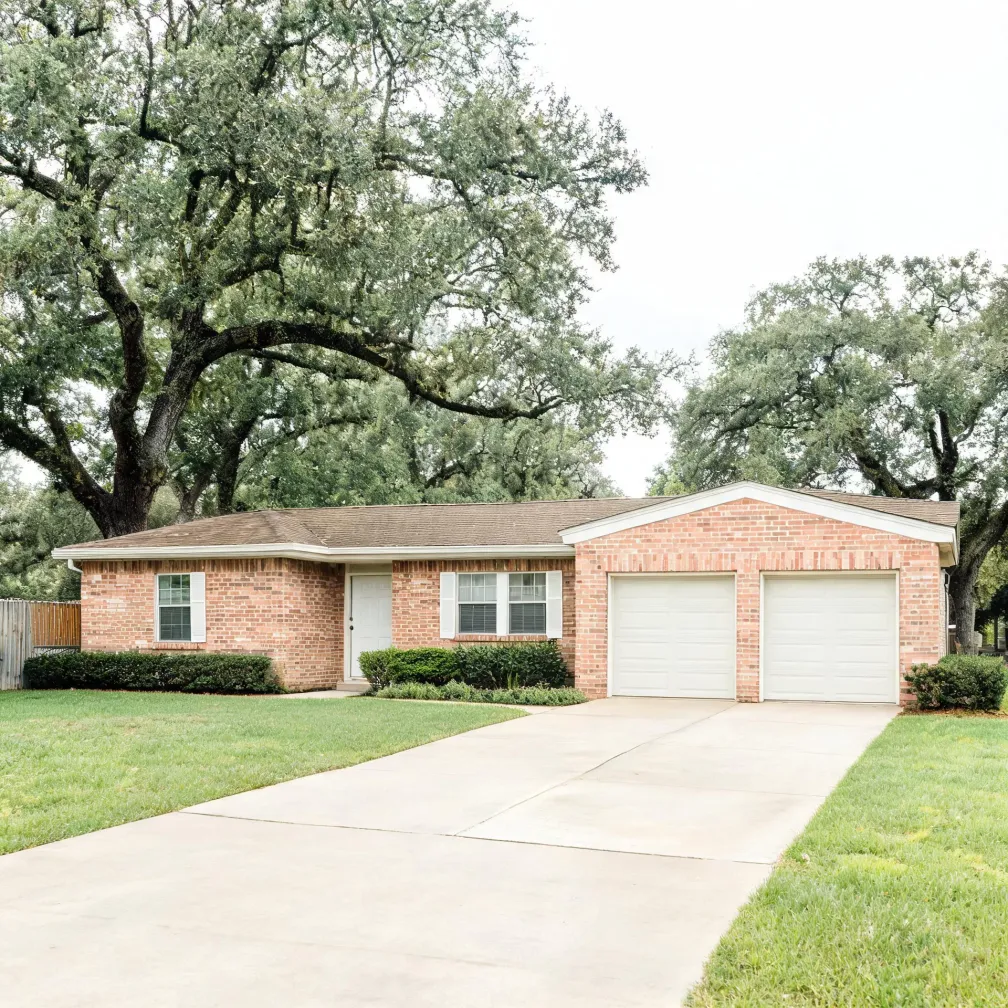 A single-story red brick house with a shingled roof, white front door, two white garage doors and a wide concrete driveway, surrounded by mowed lawn, trimmed shrubs and large oak trees under an overcast sky.
