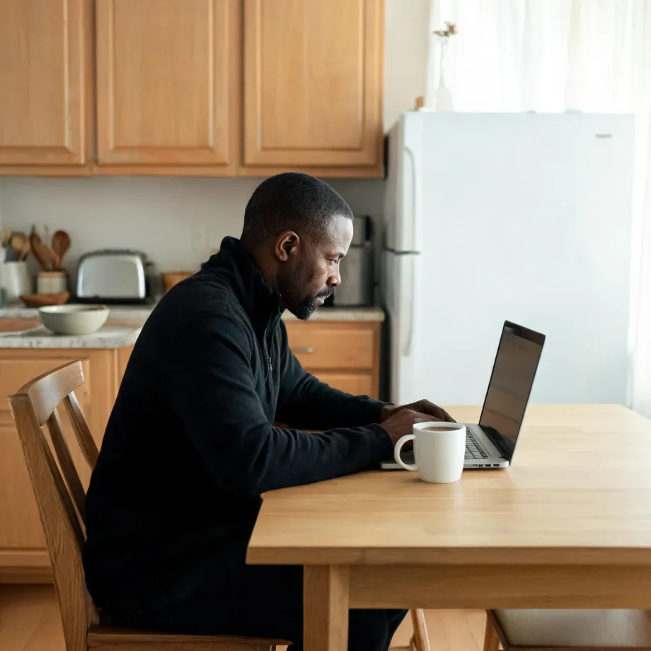 A man in a dark quarter-zip sweater sits at a wooden kitchen table using a laptop, with a white mug beside him and wood cabinets, a toaster, and a white refrigerator in the background.