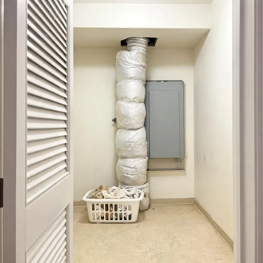 A small closet with louvered bifold doors open, showing a plastic-wrapped insulated duct running from floor to ceiling in the corner, a gray electrical panel on the wall, and a white laundry basket with cloth on the floor.