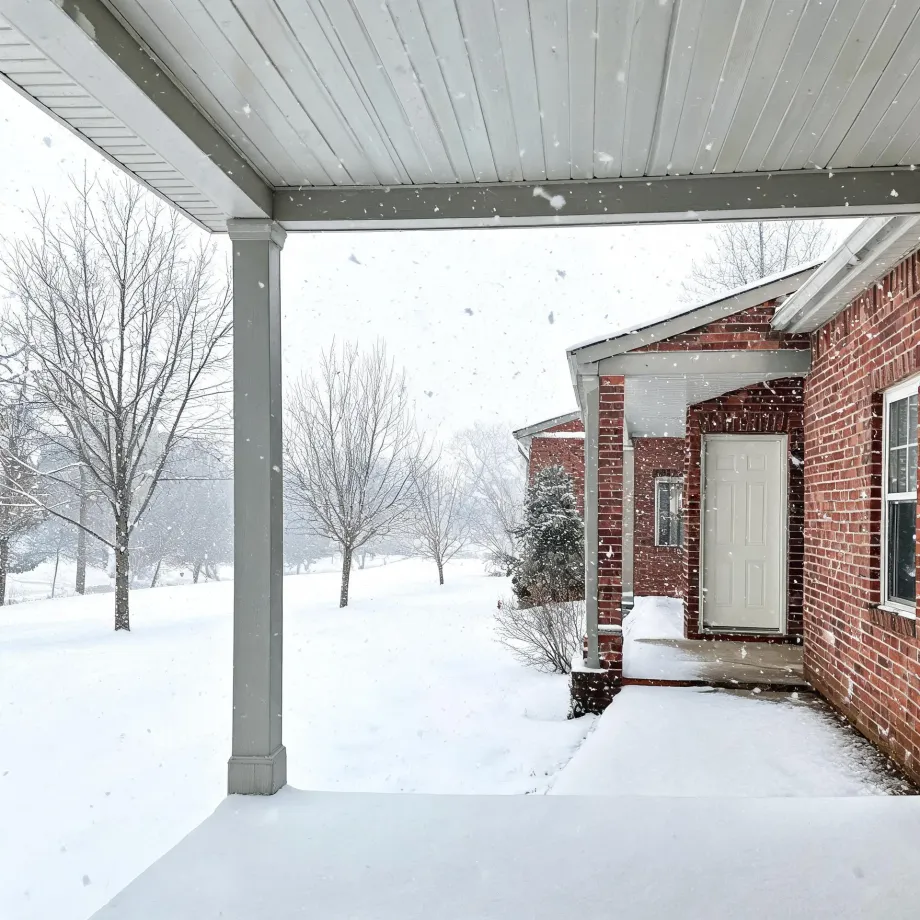 View from under a covered porch with a gray post toward a red brick house with a white door, surrounded by snow-covered ground and bare trees during falling snow.