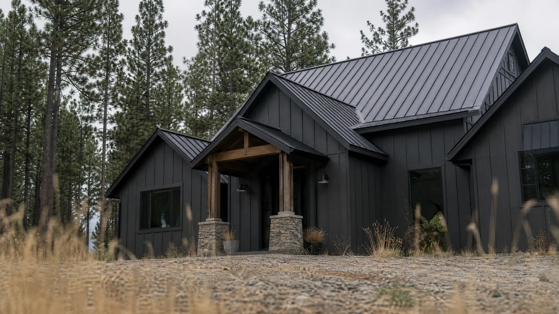 A single-story black board-and-batten house with a dark standing-seam metal roof, timber entry porch posts on stone bases, set among tall pine trees with dry grass in the foreground under an overcast sky.