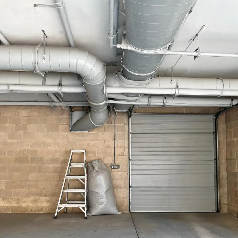 Interior of a concrete-block garage with exposed metal ventilation ducts and pipes on the ceiling, a closed roller shutter door, a folding step ladder and a filled white sack against the wall.