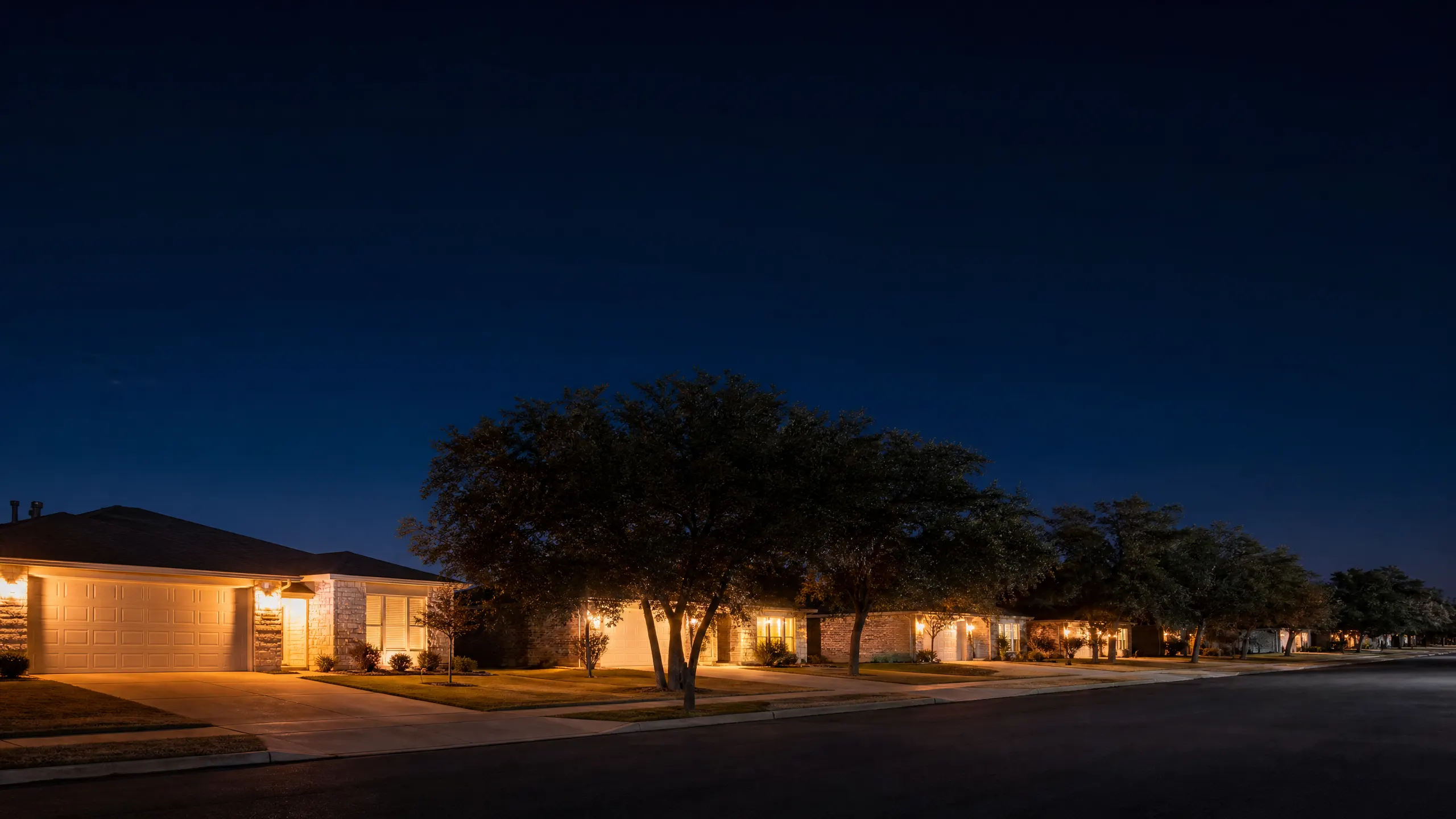 A nighttime view of a residential street lined with single-story brick houses and large trees lit by warm exterior lights.