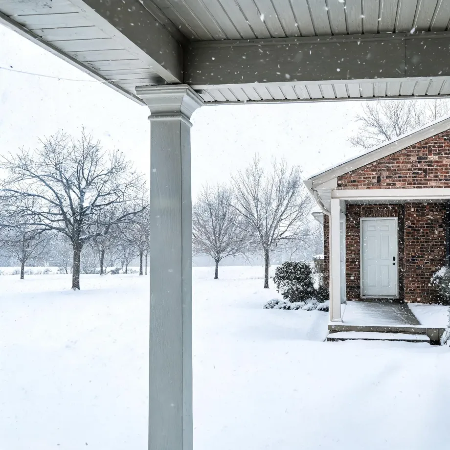 View from under a covered porch with a square white column toward a brick house with a light-colored front door, surrounded by snow-covered ground, bare trees, and falling snow.