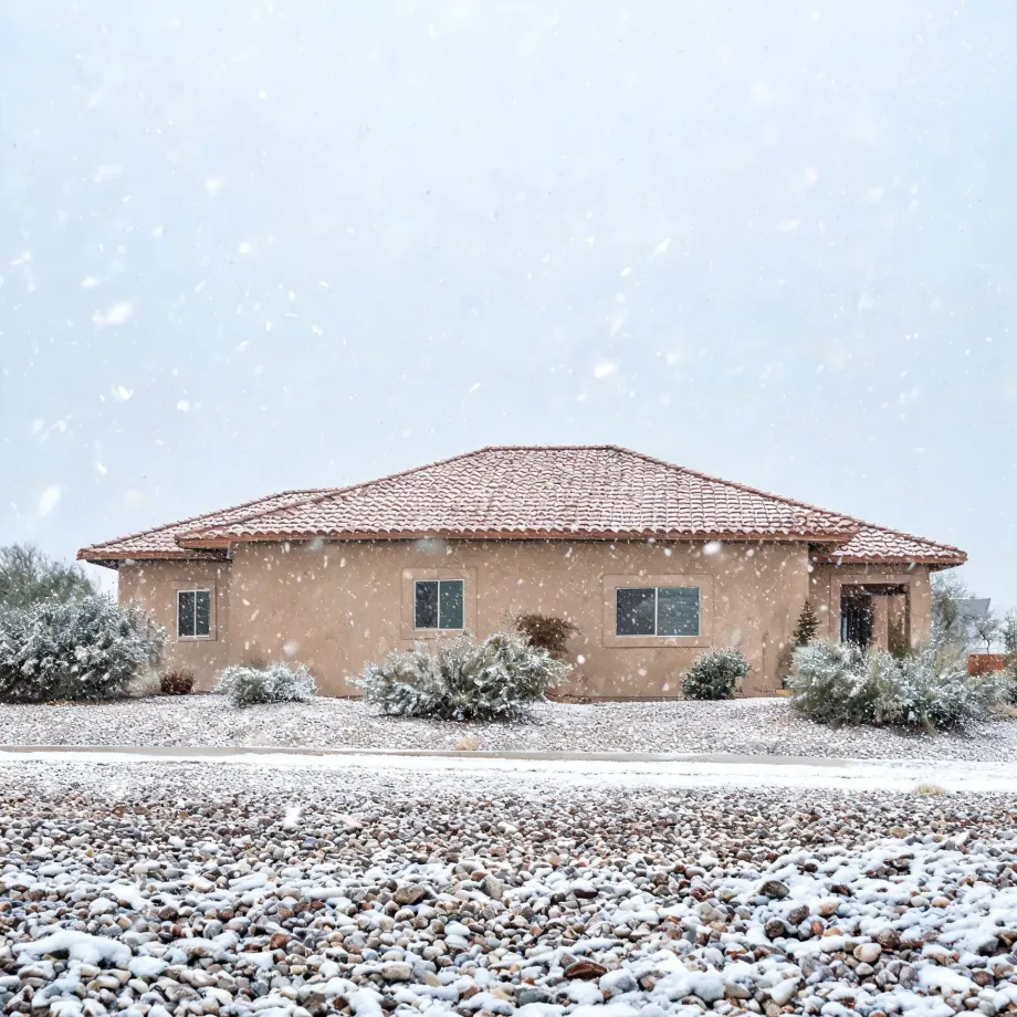 A single-story stucco house with a red tile roof during falling snow, with light snow covering shrubs and a gravel yard in the foreground.