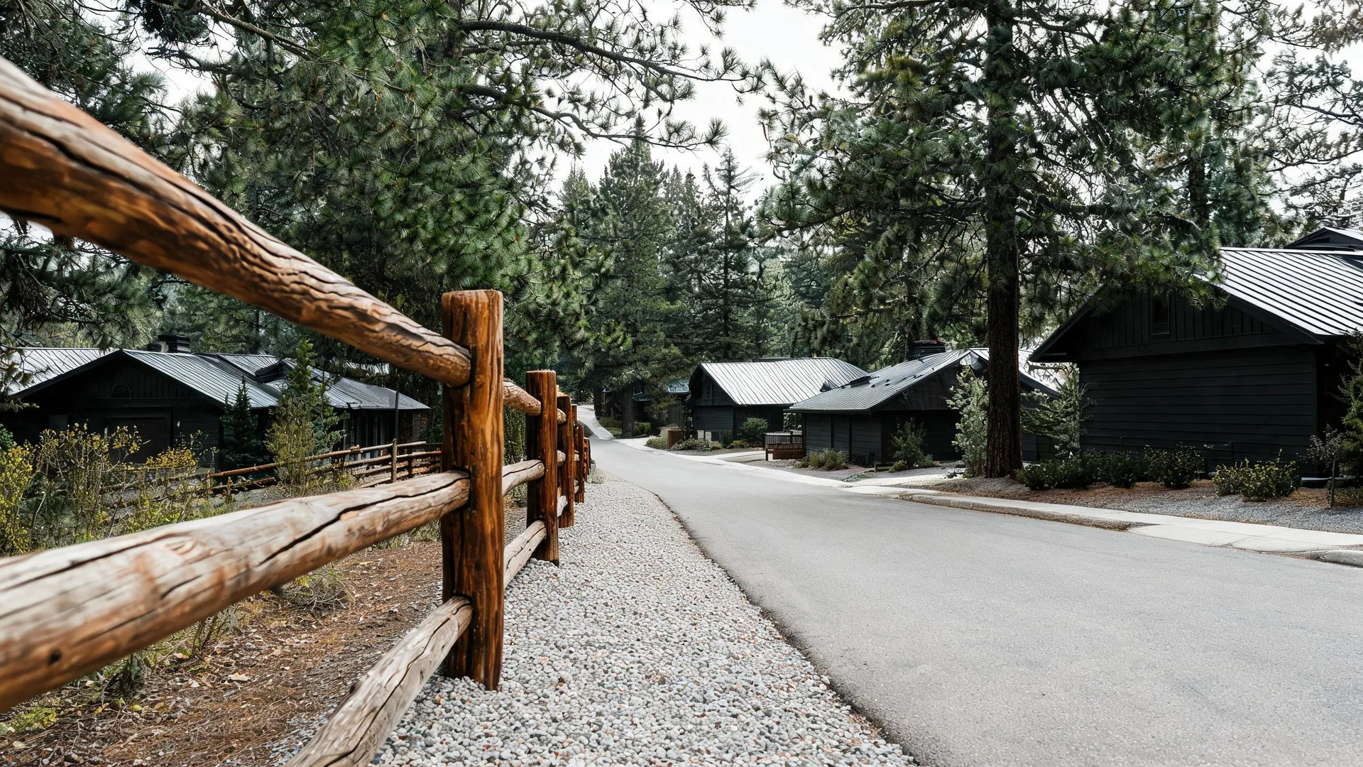 A paved road with a gravel shoulder runs past dark-clad houses with metal roofs among tall conifers, bordered in the foreground by a rustic split-log rail fence.
