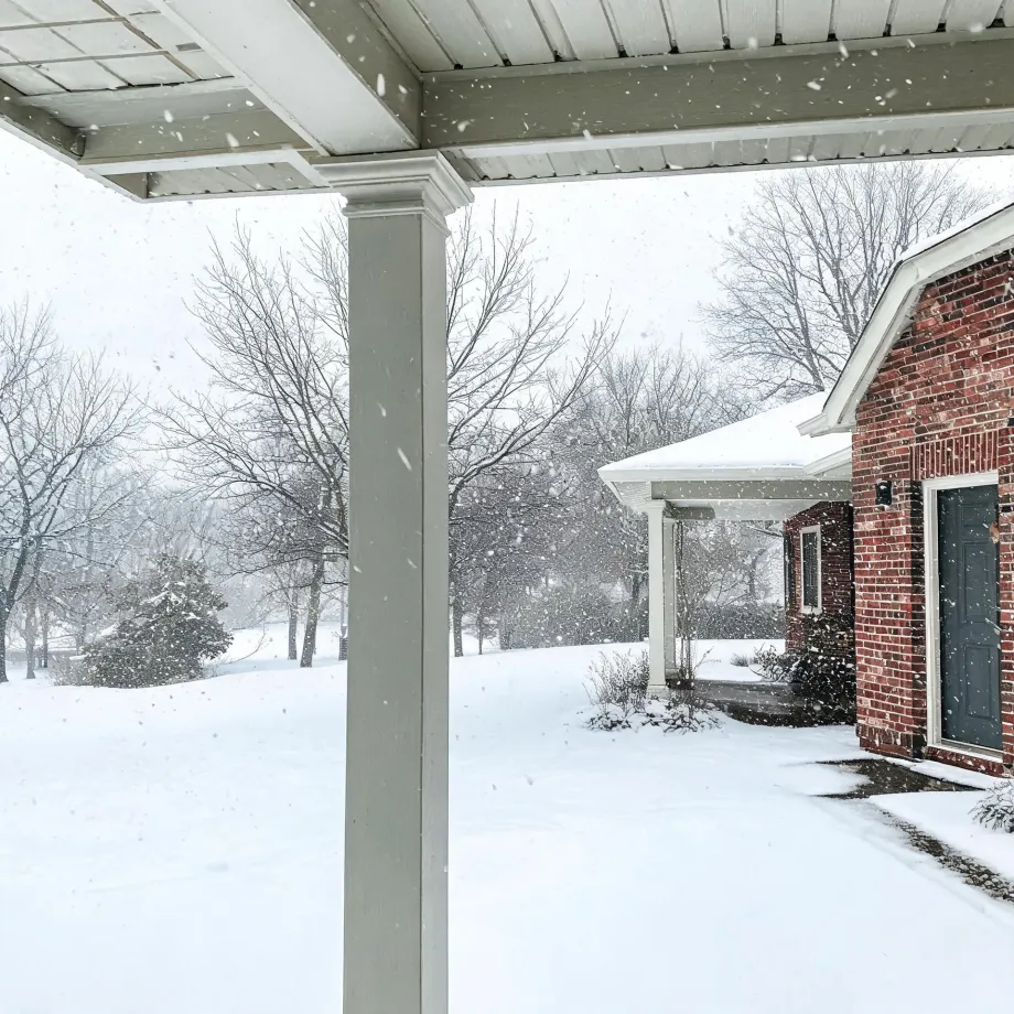 View from under a covered porch with a white square column, looking out at falling snow over a snow-covered lawn, bare trees, and a red brick house with a dark door and white-trimmed porch.