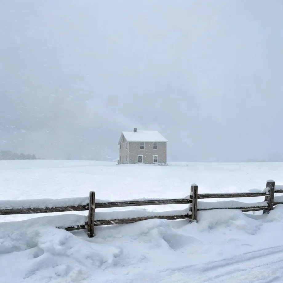 A two-story house with a snow-covered roof and chimney stands alone in a white field during falling snow, with a wooden post-and-rail fence in the foreground.