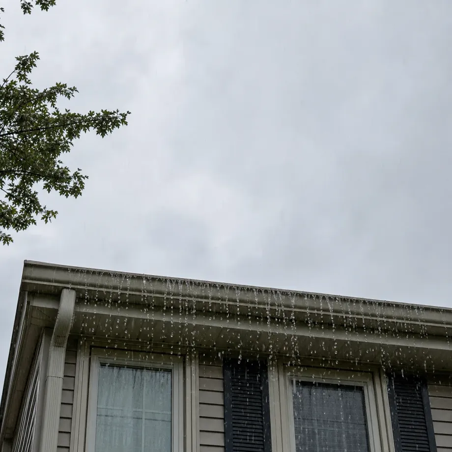 Water overflows in a sheet from the gutter along the roof edge of a house with light horizontal siding, dark shutters and windows, under an overcast grey sky with tree branches at upper left.