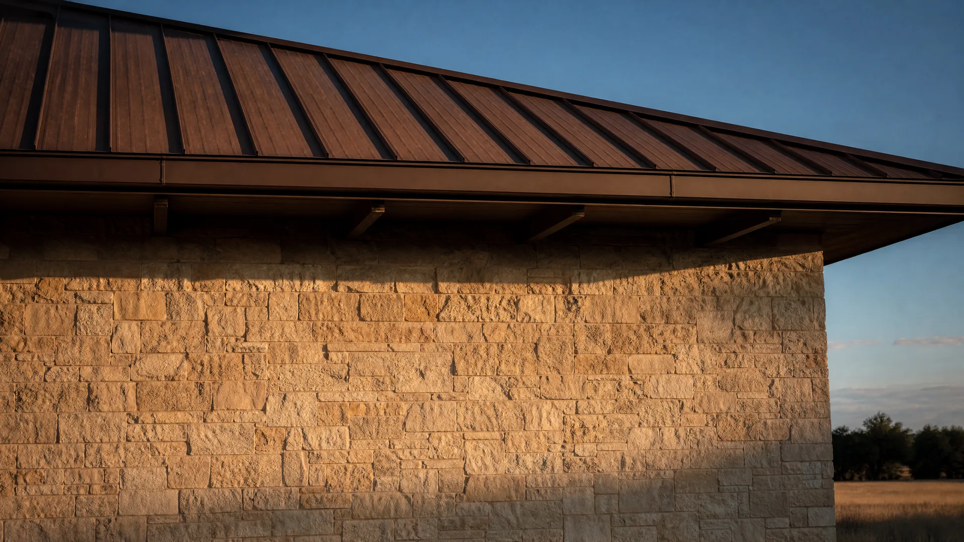 Corner of a building with a stacked limestone wall and a dark standing-seam metal roof with exposed rafter tails, lit by low warm sunlight, with blue sky and distant trees and dry grass at right.