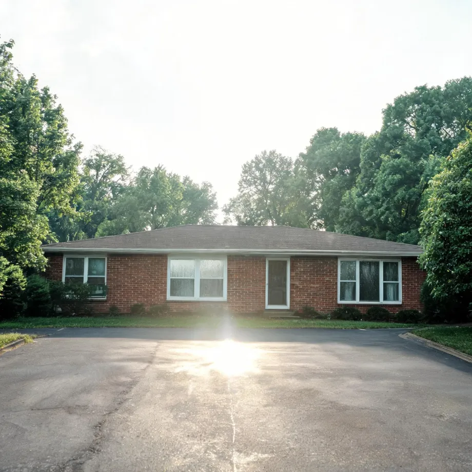 A single-story red brick ranch house with a low shingled roof, white-framed windows and a central door, fronted by a large asphalt parking area with sun glare, surrounded by leafy green trees.