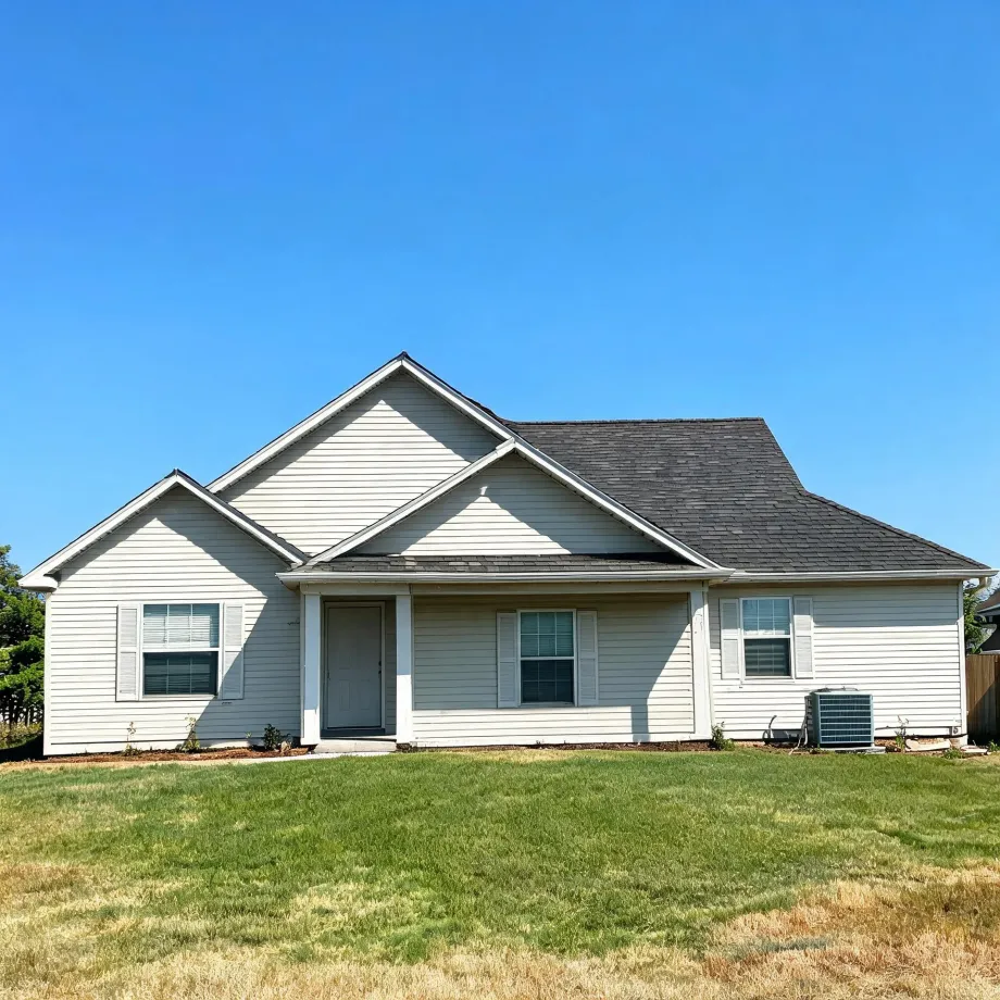 Single-story house with cream vinyl siding, gray shingled gable roof, small front porch with gray door, shuttered windows, and an outdoor AC unit, fronted by a partly dry lawn under a clear blue sky.