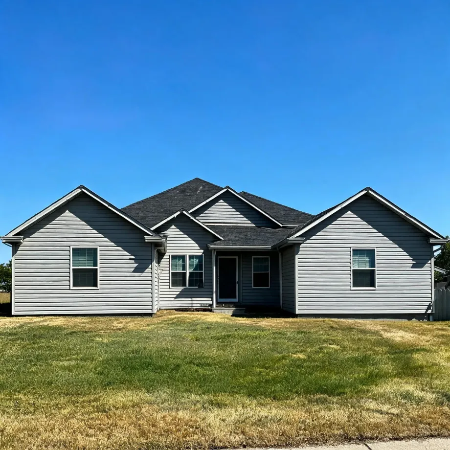 Single-story gray horizontal-siding house with dark shingled gabled roof, central entry door and several windows, fronted by a patchy green and brown lawn under a clear blue sky.