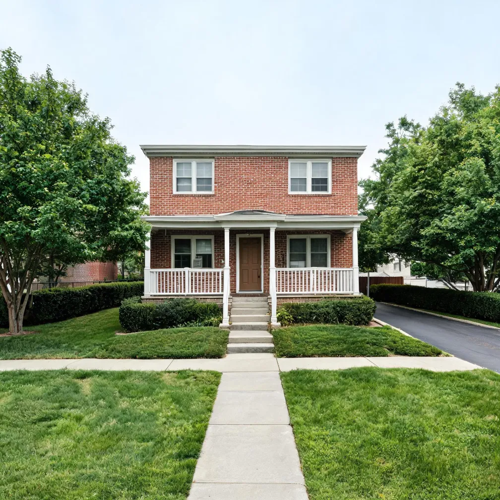Two-story red brick house with a white covered porch and railing, brown front door, and concrete walkway crossing a mown lawn, flanked by shrubs and leafy trees under an overcast sky.