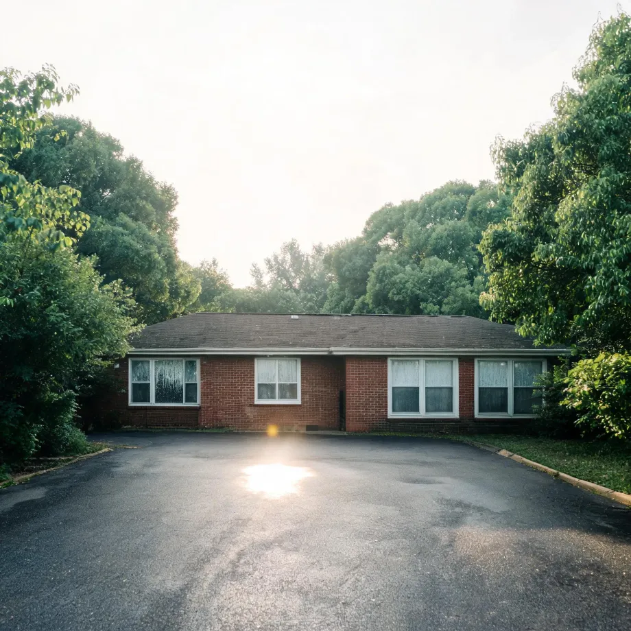A single-story red brick building with white-framed windows and a low shingled roof, fronted by a large empty asphalt parking lot with sun glare, surrounded by dense green trees under a bright overcast sky.