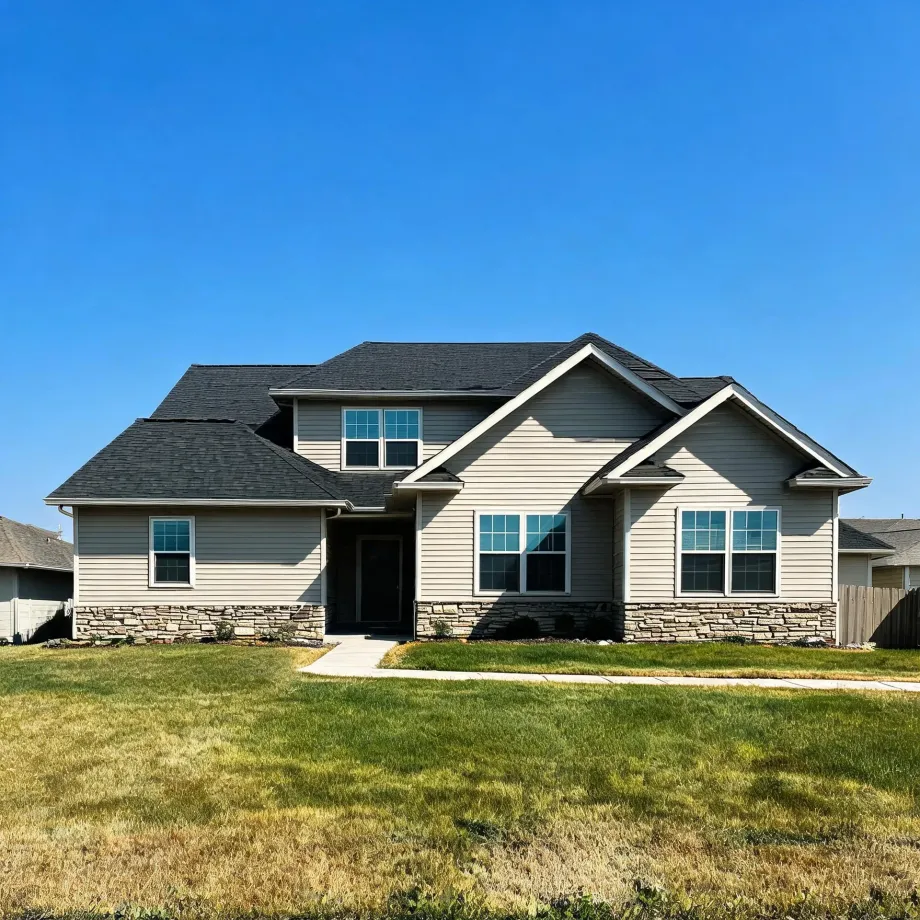 A two-story house with beige horizontal siding, stone veneer at the base, dark shingled gable roofs and a concrete walkway leading to the front door, set behind a patchy green lawn under a clear blue sky.