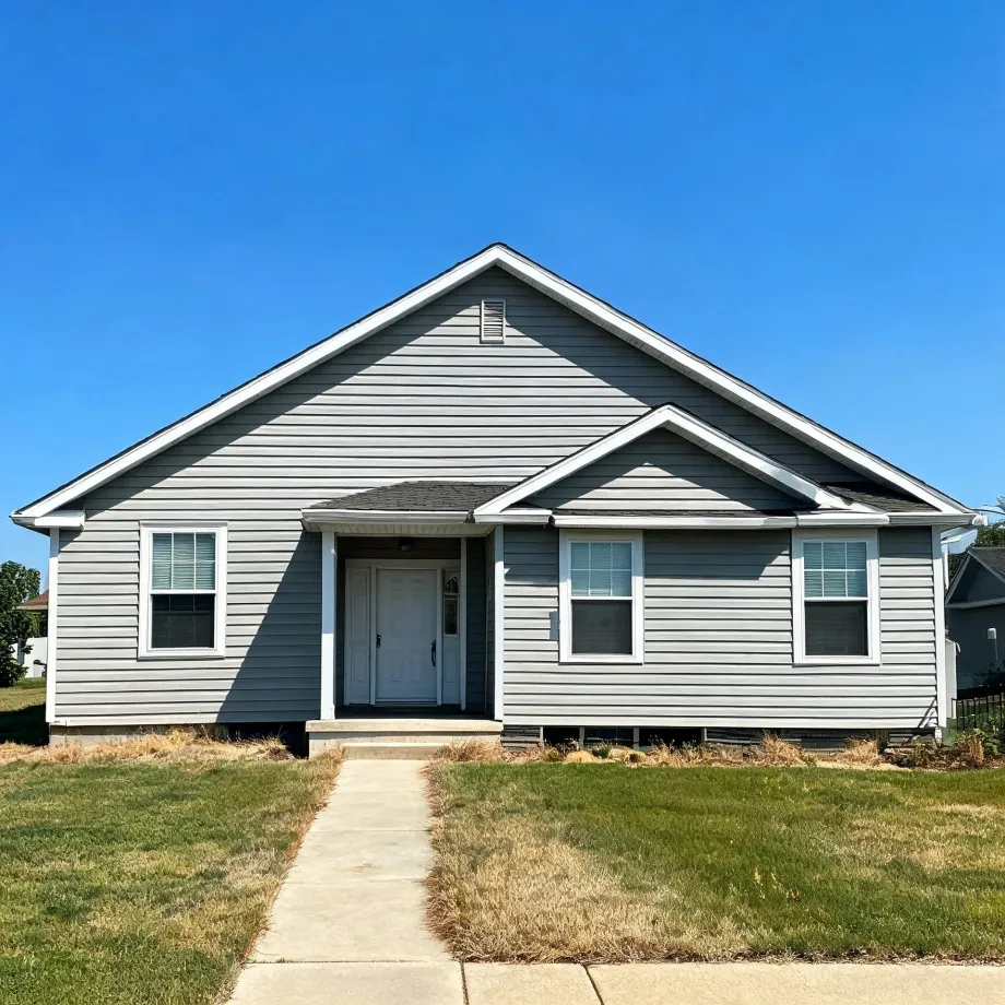 Single-story gray horizontal-siding house with white trim, gabled roof, white front door, and two windows, fronted by a concrete walkway through a patchy lawn under a clear blue sky.