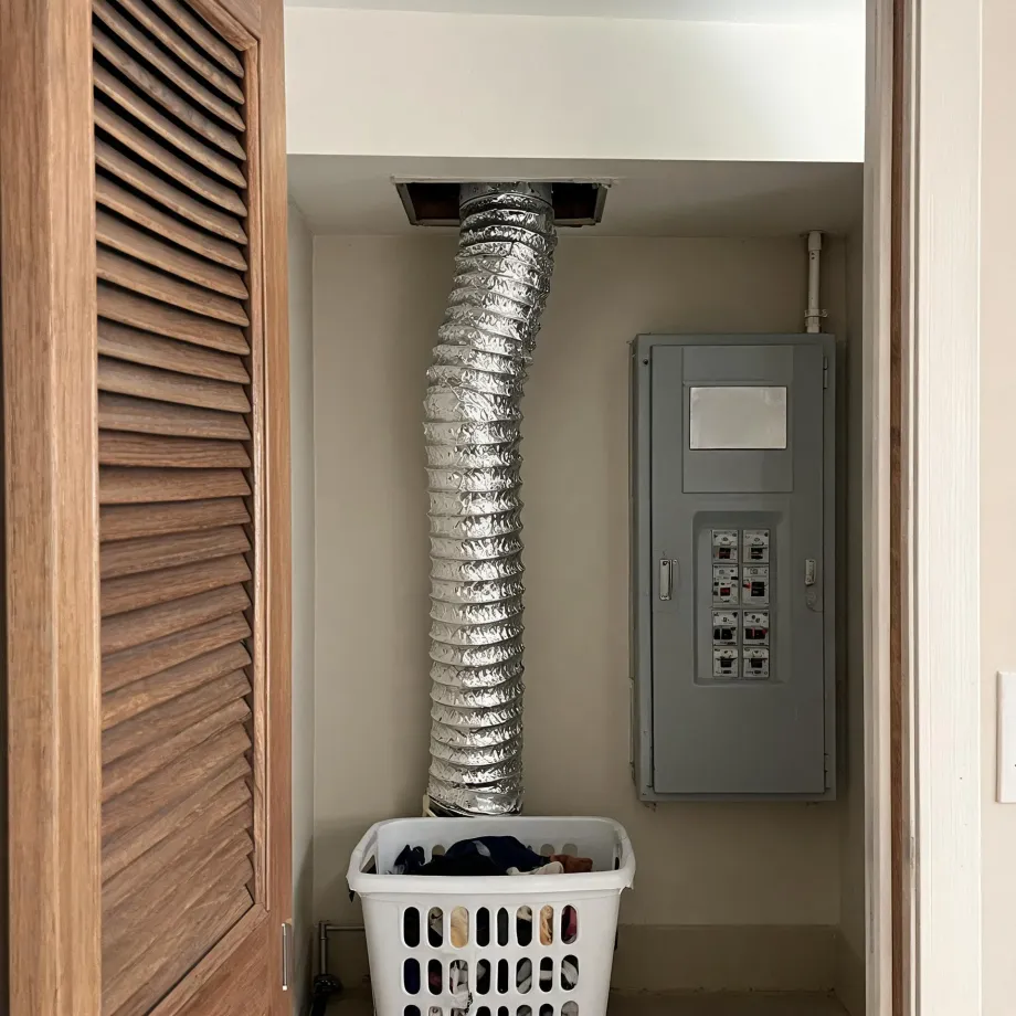 A laundry closet with louvered wooden bifold doors, a flexible foil dryer duct running from the ceiling down the back wall, a gray electrical panel on the right, and a white plastic laundry basket holding clothes on the floor.