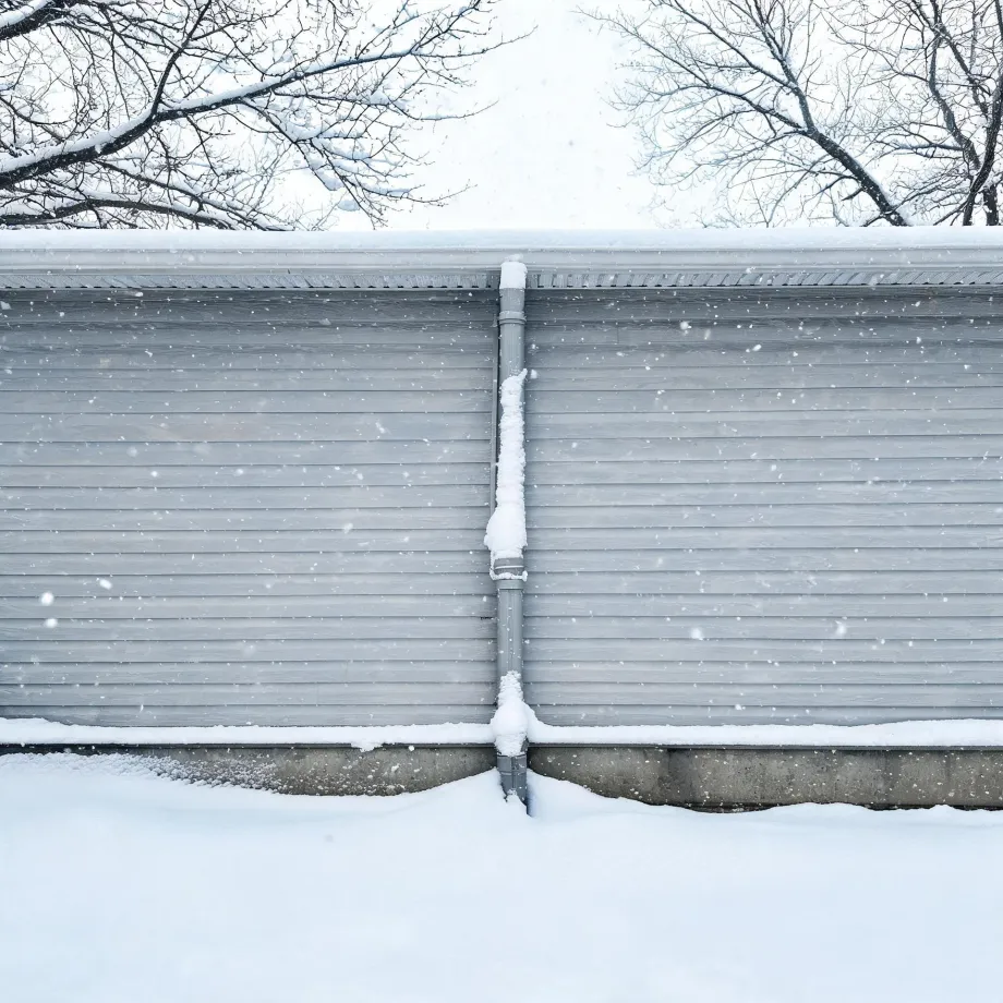 Exterior of a gray horizontal-lap-sided building with a snow-covered roof edge and a central downspout, seen during falling snow with deep snow on the ground and bare tree branches above.