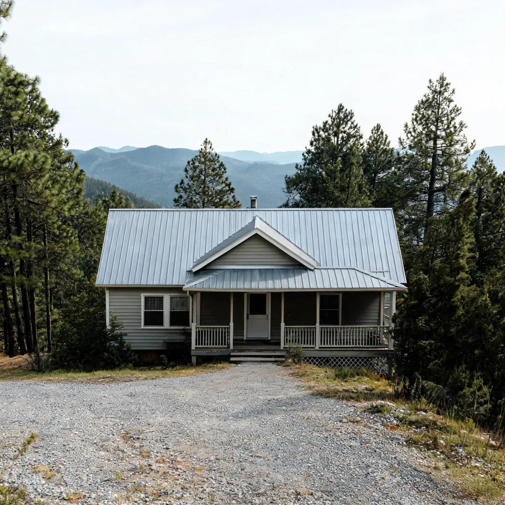 A single-story house with gray horizontal siding, a metal gable roof and a covered front porch, set at the end of a gravel driveway among tall conifers with forested mountain ridges in the background.