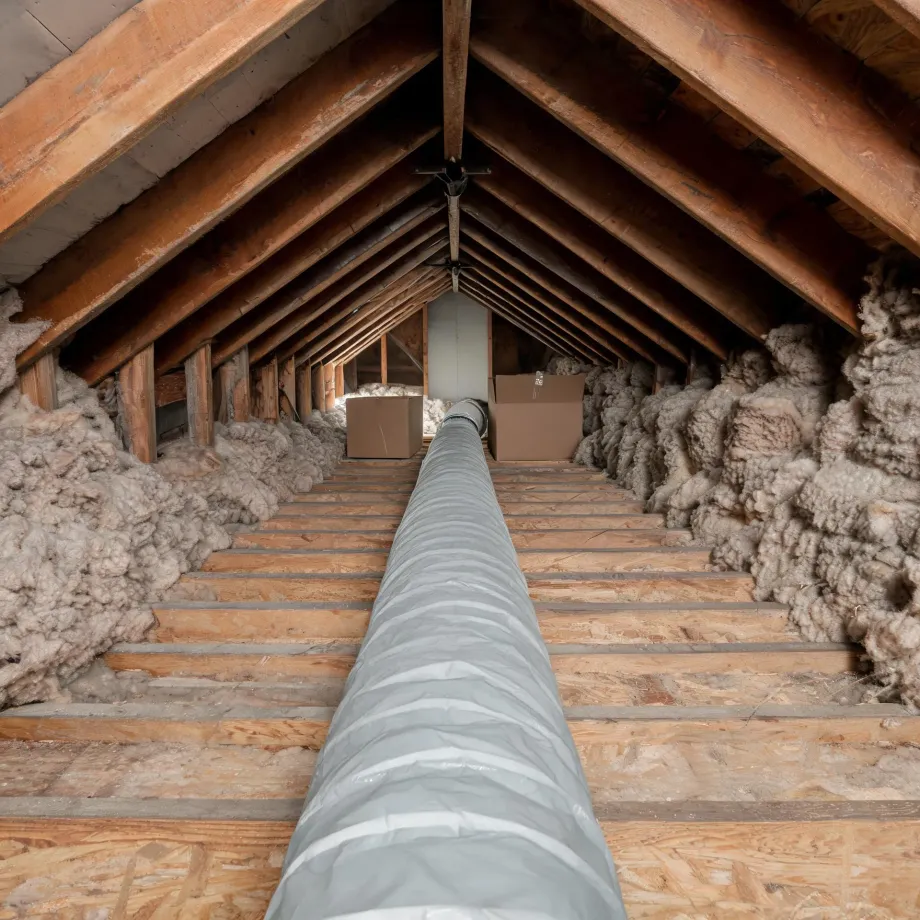 Interior of an unfinished attic with exposed wood rafters, plywood decking over floor joists, loose fiberglass insulation along both eaves, a long silver flexible duct running down the center, and two cardboard boxes at the far end.