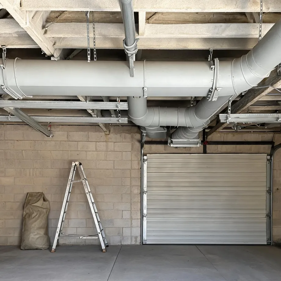 Interior of a concrete block garage with large gray ventilation ducts running along the ceiling, a closed metal roller door, an aluminum stepladder, and a canvas sack on the floor.